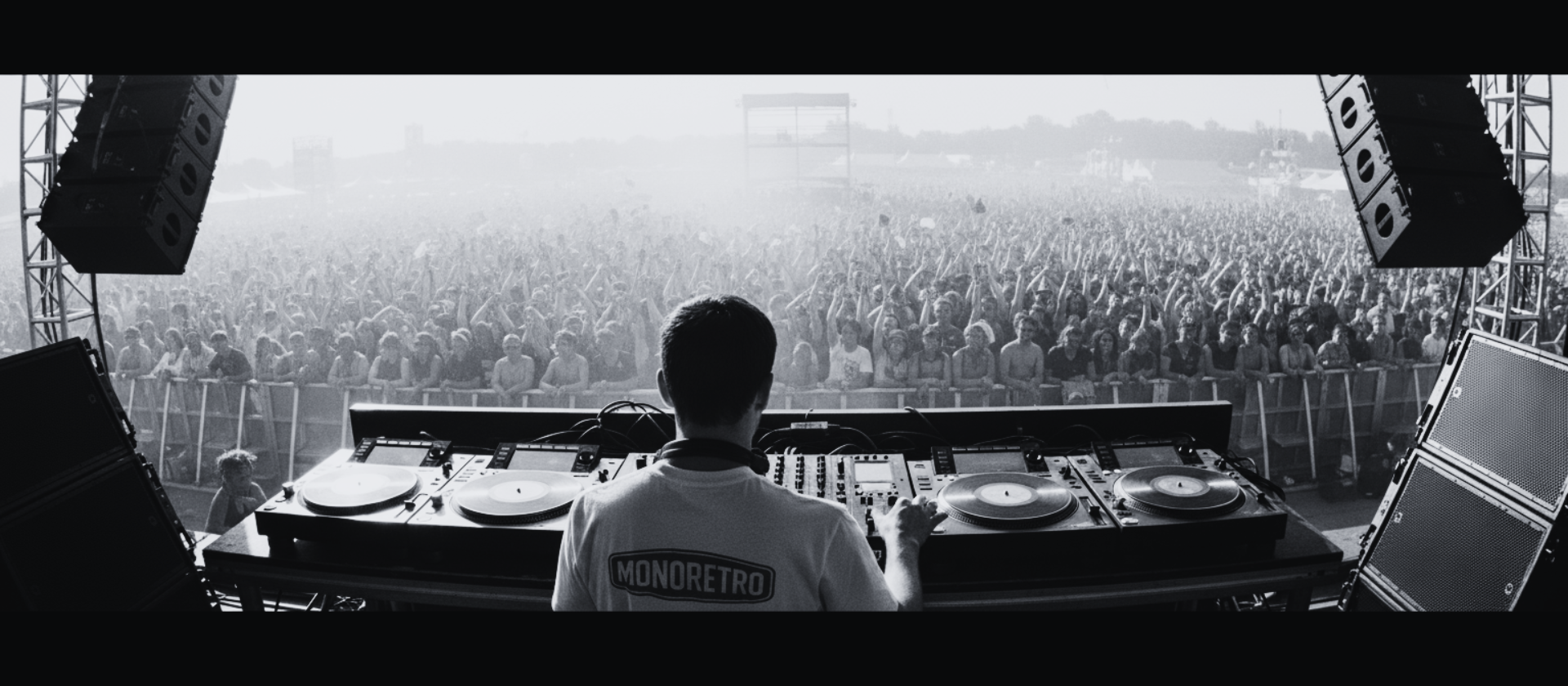 DJ in a MONORETRO T-shirt behind the decks facing a festival crowd at a beachfront stage.