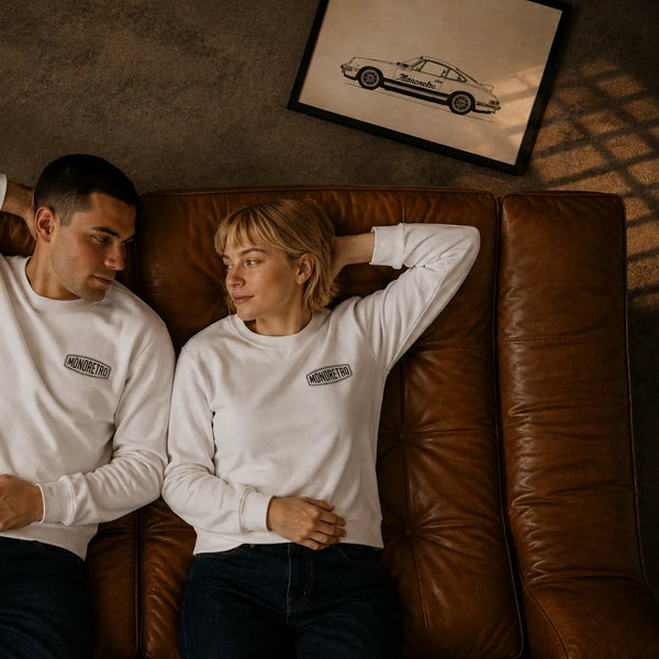 Overhead frame of two models lying on a leather sofa in white MONORETRO sweatshirts.